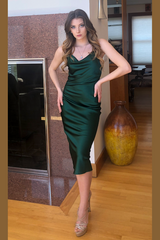 Woman in a green dress standing in a room with wooden floor and decorative vase.
