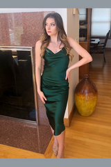 Woman in a green satin dress standing in a room with wooden floor and decorative vase.