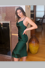 Woman in a green satin dress standing in a room with wooden floor and large vase.